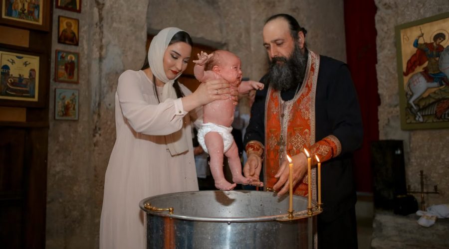 A baby being baptized by a priest and woman in a traditional church setting.