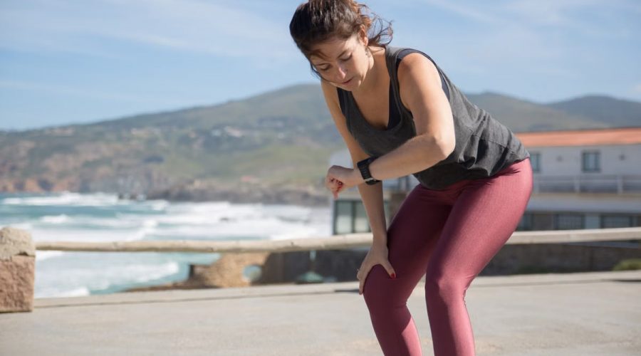 A woman in activewear checks her smartwatch while exercising outdoors by the sea.